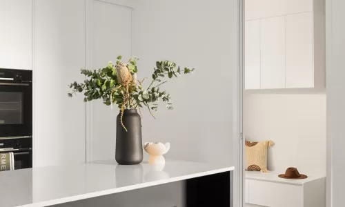 A modern kitchen with black stools, a white countertop, and a vase with green foliage, leading to a cozy room