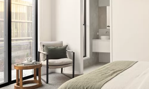 Bright bedroom with a cozy chair, wooden side table, and bed with a green throw, leading to a bathroom