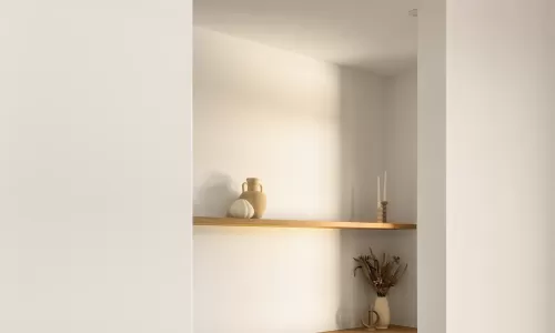 A home office with a wooden desk, grey chair, floating shelves, and minimalist decor, seen through a doorway