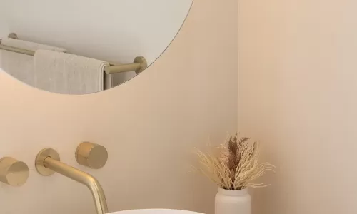 A modern bathroom with a white oval sink, gold faucet, round mirror, and decorative dried plants in a small white vase