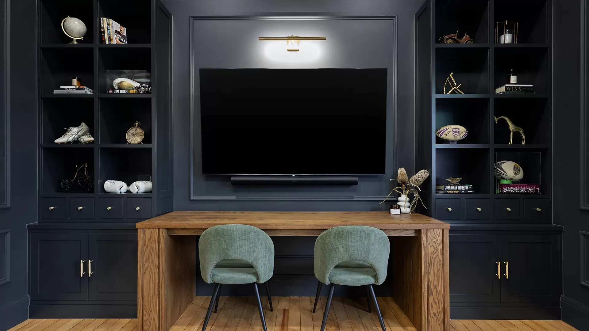 Modern office with dark built-in shelves, a TV, and two green velvet chairs.