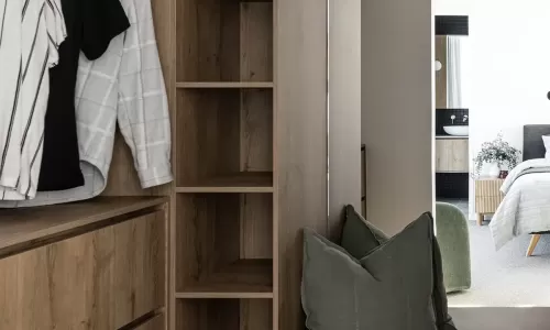 A modern walk-in closet with wood cabinetry, hanging clothes, open shelving, and a bench with green cushions