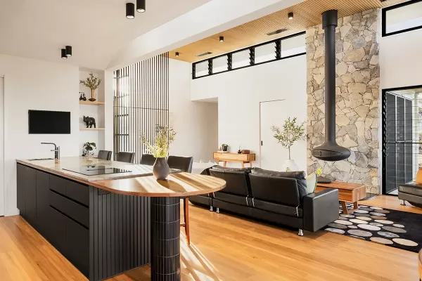 Modern open-plan living room with wooden floors, a black kitchen island with marble top, a black leather couch, a hanging fireplace, and a stone feature wall