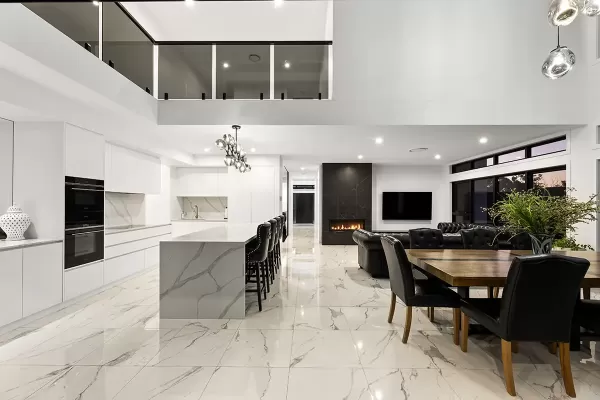 A modern open-plan kitchen and dining area with white cabinets, marble countertops, a chandelier, a fireplace and a wooden dining table with black tufted chairs