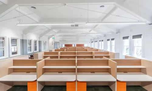 Bright open-plan office with rows of light wood workstations, white desks, and orange drawer units.