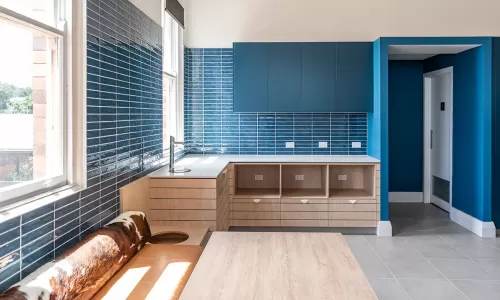 Modern kitchenette with blue tile backsplash, wood cabinetry, and a bench seat beside large windows.