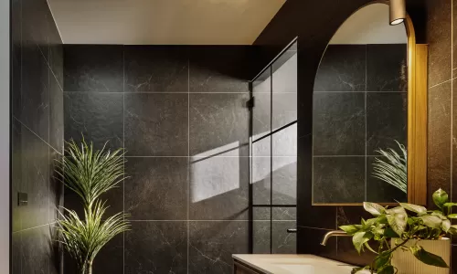 Elegant bathroom with black tile walls, wood vanity, arched mirror, and natural lighting.