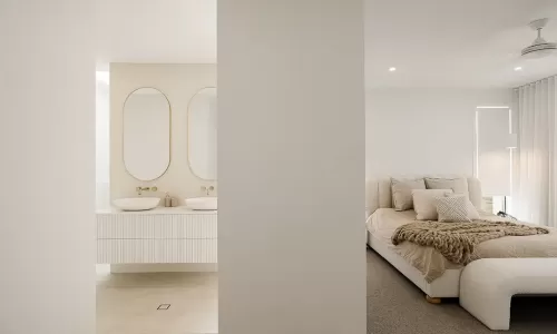 A modern bedroom with a neutral color palette and a bed with beige linens and a chunky knit throw, adjacent to an ensuite bathroom with twin oval mirrors and white vanity units