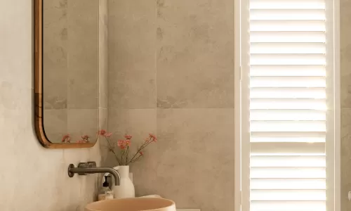 Modern bathroom with wooden vanity, round beige sink, large mirror, and shuttered window for natural light