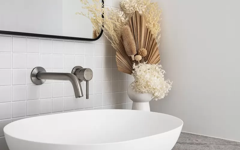 A modern bathroom vanity with a white vessel sink, gray stone countertop, white tile backsplash, and dried floral arrangement