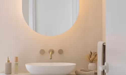 A minimalist bathroom with a round mirror, white vessel sink with golden faucet, and beige countertop with soap dispenser, small vase with dried plants, and folded towels