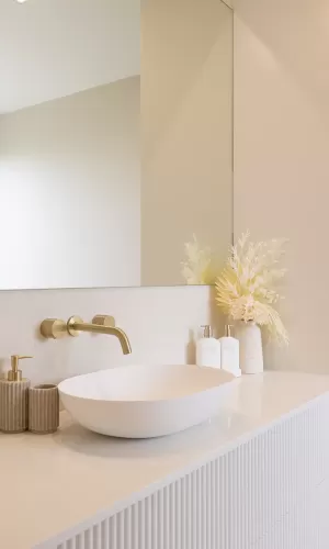 A modern bathroom with a white oval-shaped sink with a gold faucet and a vase with white and yellow dried flowers