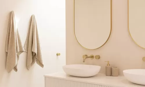 A modern bathroom with beige walls, featuring two oval-shaped mirrors with gold frames above a white vanity with ribbed drawers