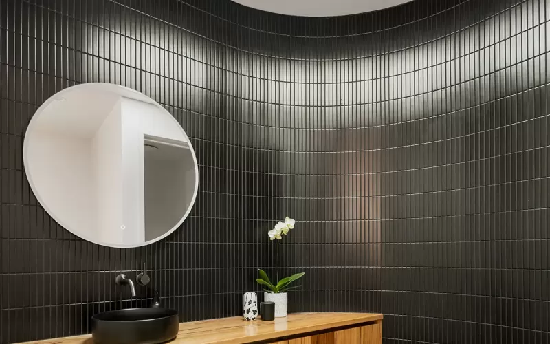 A modern bathroom with black tiled walls, a wooden countertop with built-in drawers, a round mirror, and a black sink