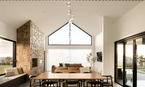 Modern living room with vaulted ceiling, stone fireplace, large windows, and a dining table with black chairs