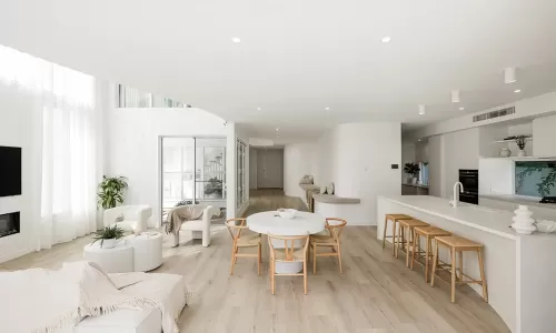 A modern, open-plan living and dining area with light wooden flooring, white walls, and large windows with sheer curtains