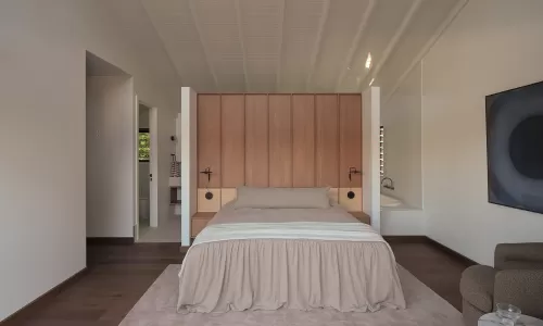 Open-concept primary bedroom with a wood-paneled headboard, neutral bedding, and a vaulted ceiling.