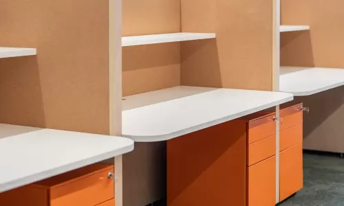 Close-up of modern office cubicles with white desks, corkboard dividers, and bright orange drawer units.