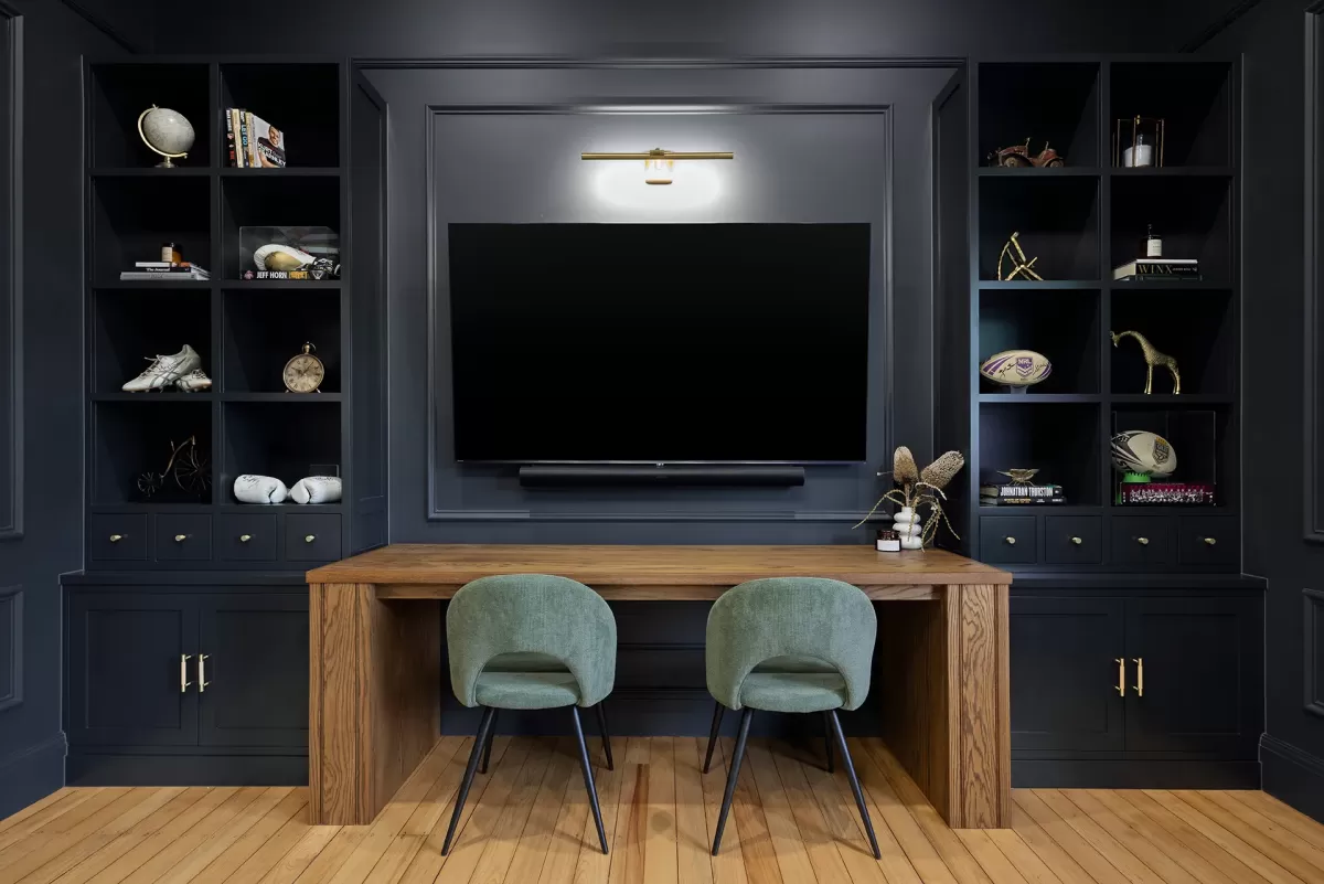 Modern office with dark built-in shelves, a TV, and two green velvet chairs.