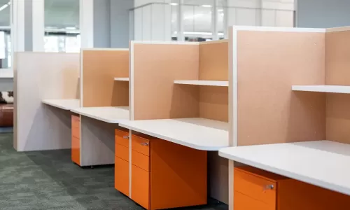 Row of clean office cubicles with corkboard dividers, white desktops, and orange filing cabinets.