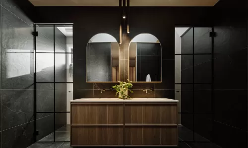 Modern bathroom with double wood vanity, arched mirrors, black tile walls, and pendant lights.