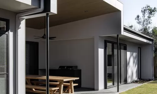 A modern outdoor patio with a wooden picnic table, barbecue grill, covered ceiling, and neatly trimmed lawn