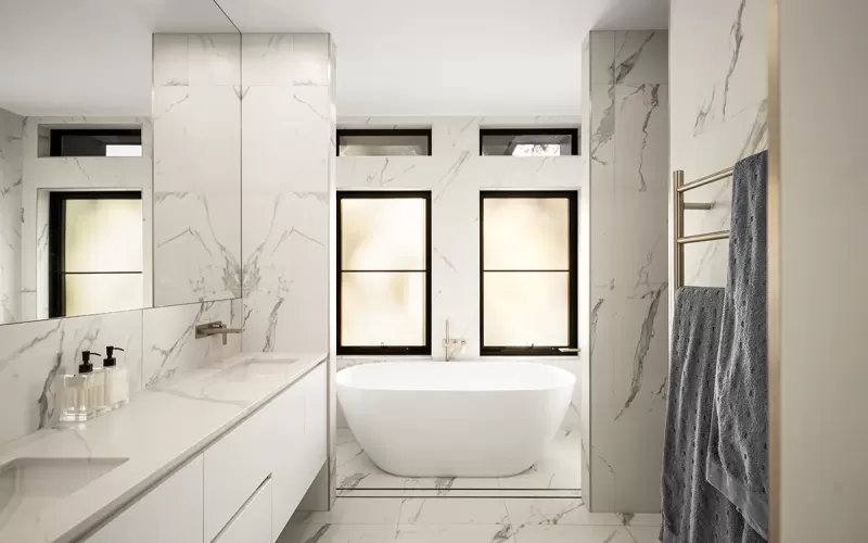 Modern bathroom with marble walls and floors, featuring a long white vanity with two sinks, a large mirror, a freestanding bathtub, and a towel rack with a grey towel hanging on it