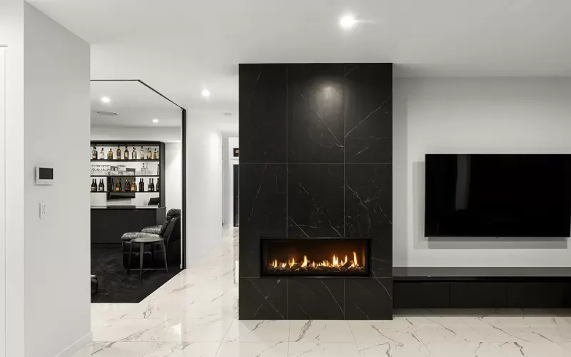 A modern living room with a black marble fireplace, flat screen TV mounted on the wall, and a home bar with bottles and glasses on the shelves in the background