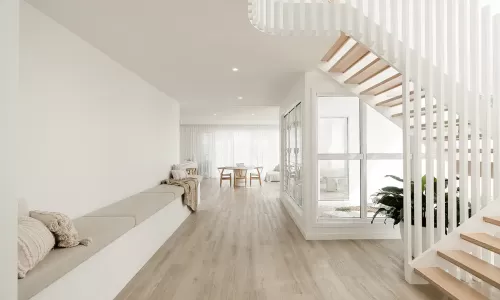 A modern, minimalist interior with light wood floors, white walls, a built-in bench with cushions, a wooden dining table with chairs, and a white staircase with vertical slats