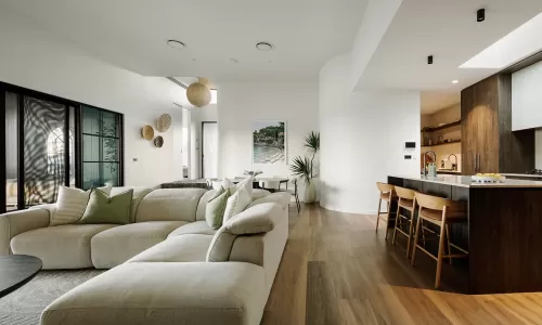Bright open-plan living area with neutral sectional, wood kitchen, and natural decorative touches.
