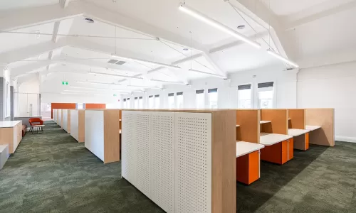 Spacious office with rows of wood-accented cubicles, orange drawer units, green carpet, and high white ceilings.
