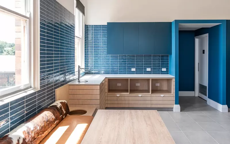 Bright kitchenette with glossy blue tile walls, light wood cabinetry, blue overhead cabinets, and a bench seat.