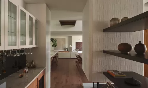 View from a wet bar down a hallway toward a living area, featuring textured tile and shelving.