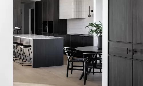 A modern kitchen with dark cabinetry, a sleek marble island, black chairs around a dining table, and light wood flooring