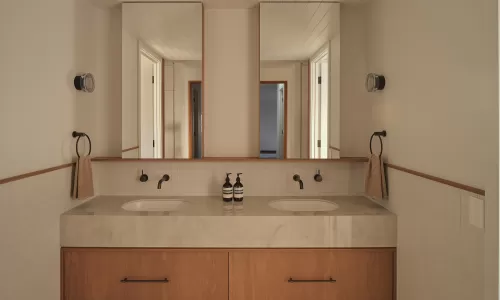 Bathroom vanity with a floating wood cabinet, double marble sinks, and two vertical mirrors.