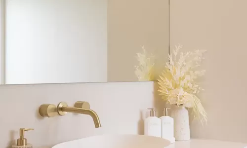 A modern bathroom with a white oval-shaped sink with a gold faucet and a vase with white and yellow dried flowers