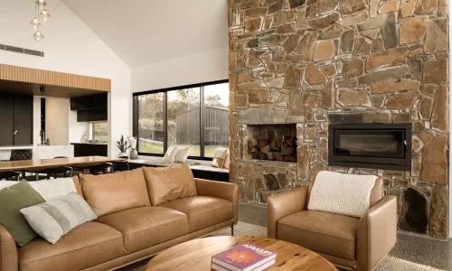 Cozy living area with stone fireplace, leather sofa, armchair, and large windows overlooking the yard