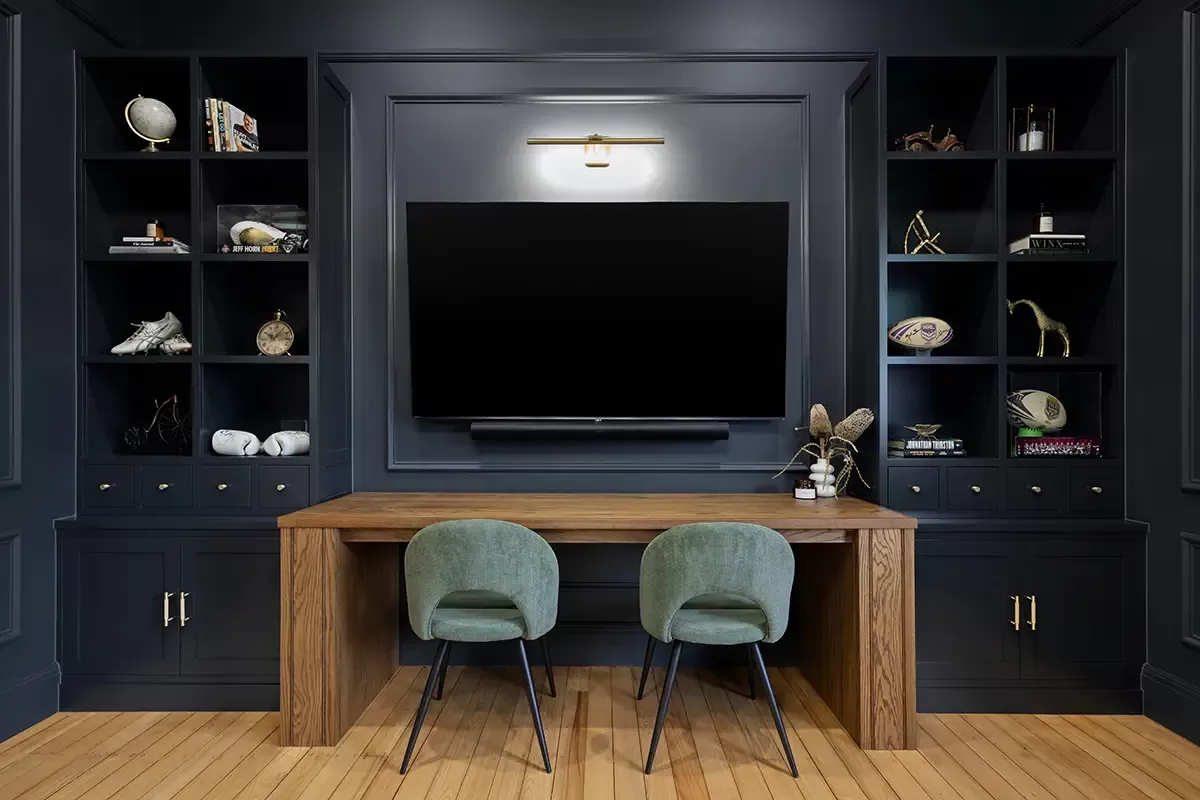 Wood desk with two green chairs facing mounted TV and shelves.
