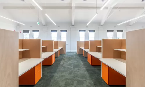 Bright office with rows of wooden cubicles, orange drawers, and large windows letting in daylight.