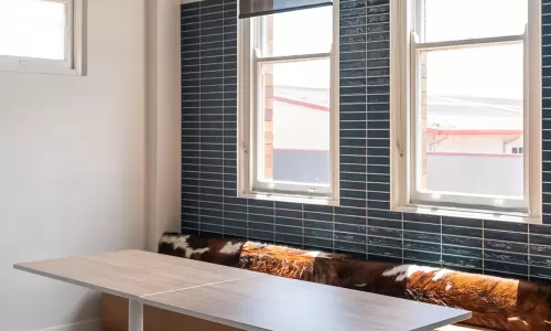 Minimalist meeting area with a long wood table, tan bench seating, and large windows with blinds.