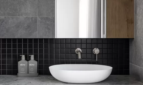 A modern bathroom with gray stone walls, a wood vanity, a round white vessel sink, and black square tiles