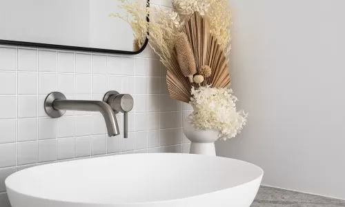 A modern bathroom vanity with a white vessel sink, a gray stone countertop, white tiles, and dried floral decor