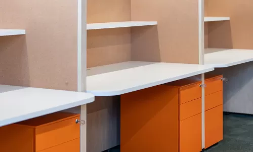 Office cubicles with white desktops, corkboard partitions, and bright orange mobile pedestal drawers.